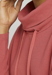 Bluza z kapturem w kolorze przygaszonego różu o fakturowanej tkaninie, z wysokim kołnierzem, sznurkami i luźnym krojem. Zbliżenie na dekolt i dłoń.