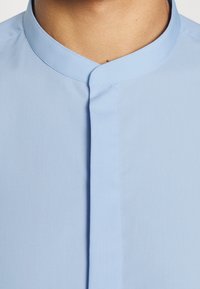 Lichtblauwe, shirt met korte kraag, met een gladde structuur en een voorpande met verborgen knopen. Eenvoudig, strak ontwerp.