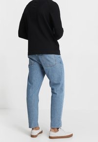 Osoba oblečená v světle modrých džínách, černém topu s dlouhým rukávem a bílých teniskách s hnědými podrážkami, stojící na pozadí v jednolitém světlém odstínu.