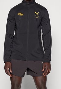 Chaqueta atlética ligera negra con cierre frontal, puños elásticos y acentos de logo amarillo en el pecho y los hombros. Textura suave.