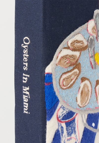 Ράχη βιβλίου με την επιγραφή «Oysters In Miami» και διακοσμητικό κέντημα με πιάτο στρείδια που απεικονίζει κοχύλια και λεμόνι σε μπλε ύφασμα.