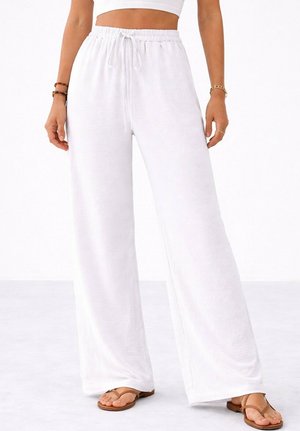 Femme portant un pantalon blanc à jambes larges avec ceinture à cordon, un crop top blanc, des sandales marron et des bracelets aux poignets.