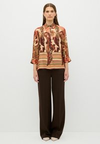 Chemisier à manches longues présentant un motif floral et rayé aux teintes de rouge, vert et beige, associé à un pantalon large marron.