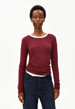 Femme aux cheveux courts et naturels portant un haut à manches longues bordeaux par-dessus une chemise blanche et un jean bleu foncé, debout devant un fond blanc.