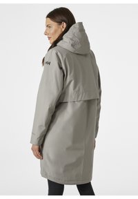 Abrigo impermeable gris con capucha, mangas largas y un corte holgado. Detalles destacados incluyen un logo en el hombro y una abertura en la espalda.
