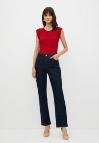 Červený žebrovaný top bez rukávů, tmavě modré džíny s vysokým pasem a béžové lodičky. Žádné vzory ani výrazné kovové detaily nejsou viditelné. Jednoduchý design.