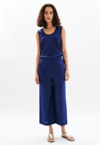 Marine blauwe mouwloze top met een ronde hals, gecombineerd met vloeiende marine blauwe wijde broek. Sandalen met een metalen accent maken de outfit compleet.