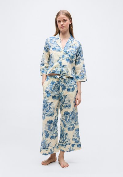 Γυναίκα που στέκεται ξυπόλυτη, φορώντας κρεμ πιτζάμες με μπλε λουλουδένια και τοπία μοτίβα, πουκάμισο με τρία τέταρτα κουμπιά και φαρδιά παντελόνια.