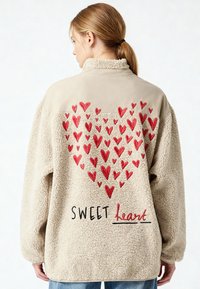 Giacca in pile beige chiaro con cuori rossi ricamati che formano una grande forma a cuore e la scritta "SWEET heart" sul retro, indossata da una persona.