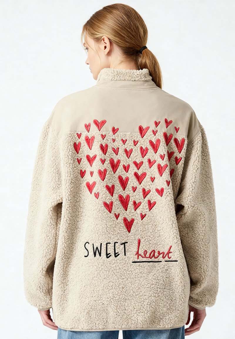 Giacca in pile beige chiaro con cuori rossi ricamati che formano una grande forma a cuore e la scritta "SWEET heart" sul retro, indossata da una persona.
