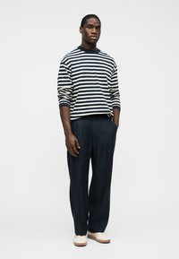 Άνδρας που στέκεται με τα χέρια στις τσέπες, φορώντας μακρυμάνικο πουκάμισο με μαύρες και λευκές ρίγες, σκουρόχρωμο παντελόνι και ανοιχτόχρωμα αθλητικά παπούτσια σε λευκό φόντο.