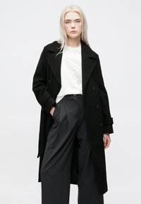 Cappotto doppiopetto nero con ampi revers, realizzato in un materiale morbido, indossato sopra una camicia bianca e pantaloni grigi a palazzo con vita alta.