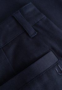 Detail na námornícko modrej látke nohavíc s pásom, zdobným šitím a plochou vreckom s embosovaným kovovým prvkom.