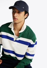 Zielono-biała, pasiasta koszulka rugby z niebieskimi akcentami, z kołnierzykiem i trzema guzikami. Ciemna czapka dodaje kontrastującego akcentu.