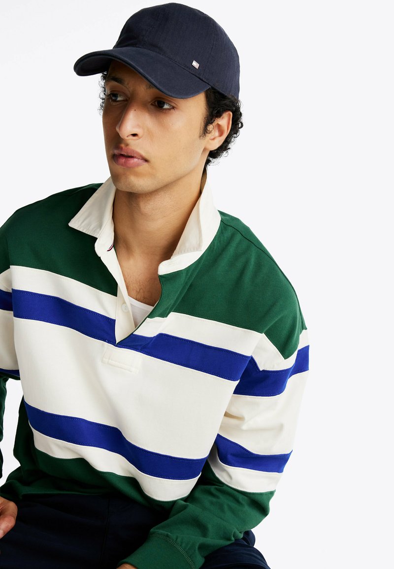 Zielono-biała, pasiasta koszulka rugby z niebieskimi akcentami, z kołnierzykiem i trzema guzikami. Ciemna czapka dodaje kontrastującego akcentu.