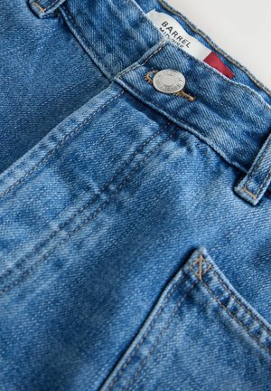 Modré džínové kalhoty z blízka zobrazující knoflík, poutko na opasek, šití a částečnou přední kapsu s viditelnou značkou uvnitř pasu.