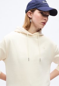 Νεαρό άτομο με κρεμ φούτερ με κουκούλα και μπλε σκουρό καπέλο, που φοράει ασημένιο σκουλαρίκι σε σχήμα σταγόνας, κοιτάζει ελαφρώς δεξιά με τα χέρια στη μέση.