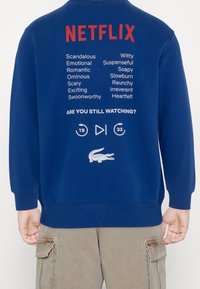 Modrá mikina s nápisem "NETFLIX" červeně na zádech, s textem v bílé barvě. Obsahuje logo krokodýla a ikony přehrávání ve spodní části.