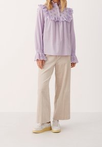 Blouse lilas à manches longues avec col volanté en broderie anglaise, manches évasées, associée à un pantalon large en velours côtelé beige et des baskets blanches.