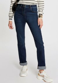 Jeans de tiro alto azul oscuro con puños enrollados, con costuras en contraste y un diseño clásico de cinco bolsillos, combinados con zapatillas blancas.