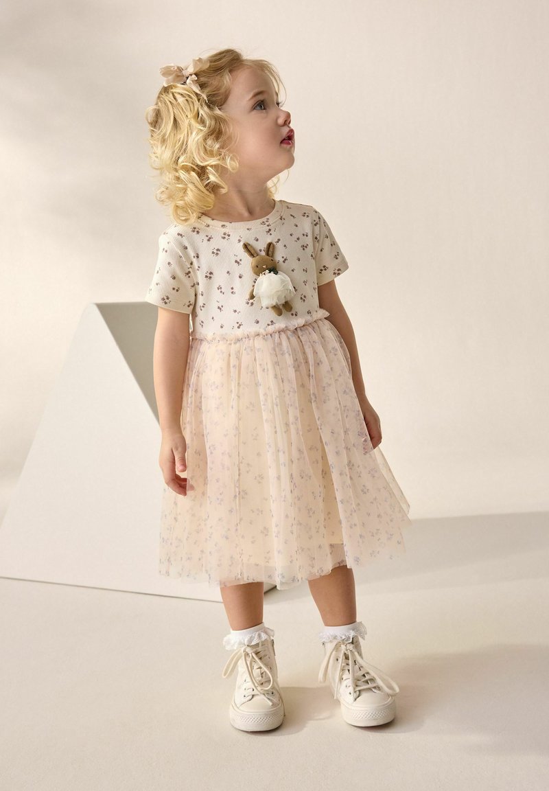 Toddler girl with curly blonde hair wears a floral dress with a bunny decoration, white socks, and lace-up sneakers, looking to the side.