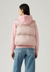 Donzen vest in lichtroze met een aanliggend boord en een capuchon, gedragen over een roze sweatshirt. Blauwe denim jeans met een losse pasvorm maken de look compleet.