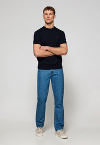 Marinblå kortärmad skjorta med ribbad halsringning, röd logotypaccent. Blå rakbenade jeans med ljus tvätt. Beiga sneakers fulländar outfiten.