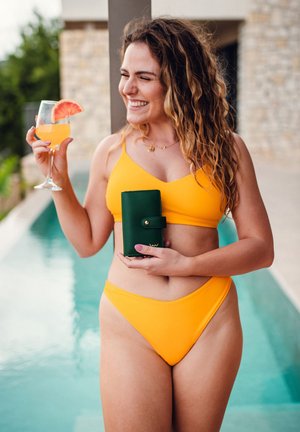 Yellow bikini and high-waisted bottoms, holding a green wallet and a glass of orange drink with a grapefruit slice, by a poolside.