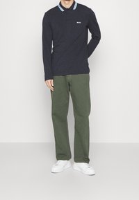 Námořnický polo shirt s dlouhým rukávem, vícebarevným límcem a bílým logem "BOSS", spárovaný se zelenými kalhotami a bílými teniskami.