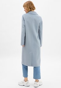 Cappotto lungo azzurro chiaro con colletto a sfondo e spacco singolo, abbinato a jeans corti e scarpe bianche, visto da dietro.