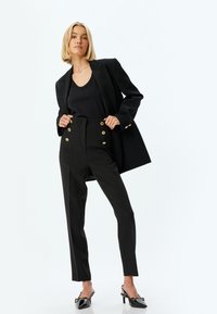 Blazer nero su misura sopra un top nero, abbinato a pantaloni neri a vita alta con dettagli in oro e eleganti tacchi neri.