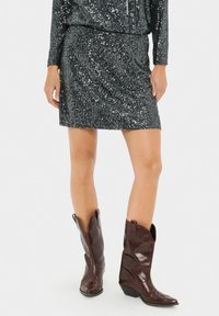Mujer vestida con una mini falda negra de lentejuelas y botas de vaquero de cuero marrón oscuro, de pie frente a un fondo blanco liso.
