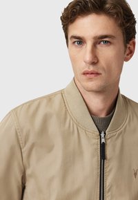 Chaqueta bomber beige con cierre de cremallera, cuello acanalado y tejido texturizado. Presenta un sutil detalle de logo en el lado izquierdo del pecho.