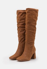 Par de botas altas de ante marrón, de caña baja, con punta afilada y tacones de bloque sobre fondo blanco.