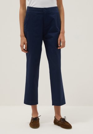 Donna che indossa pantaloni blu navy corti a gamba dritta, top bianco e scarpe da barca marroni, in piedi su uno sfondo chiaro e uniforme.