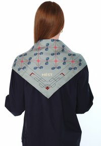Silkesjal i lyseblå med geometriske mønstre i mørkeblått og rødt; har teksten "HÉST" i gull. Klassisk firkantet form.