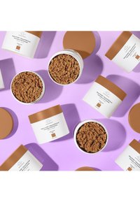 Weiße Behälter mit braunen Deckeln, die einen braun strukturierten Peeling enthalten, auf einem pastelllila Hintergrund, mit Produktetiketten, die nach oben zeigen.