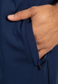 Námořnické modré sportovní kalhoty mají boční kapsu na zip s hladkou texturou, úzký střih a ruku částečně vloženou do kapsy.