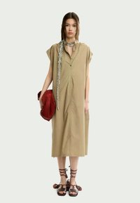 Khaki mid-length dress with short sleeves, button front, two side pockets, and a relaxed fit. Worn with strappy sandals and a red bag.