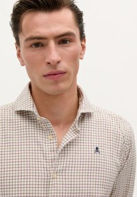Button-up Hemd mit Karomuster in Creme, Lila und Marineblau, mit einem marineblauen Totenkopf-und-Kreuzsch bones Logo auf der Brust.