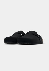 Μαύρα ξώφτερνα με λεία, ματ επιφάνεια. Διαθέτουν λουράκι με αγκράφα, στρογγυλεμένη μύτη και υφή στην εξωτερική σόλα για καλύτερη πρόσφυση.