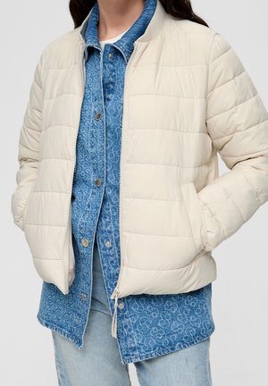 Vrouw draagt een crèmekleurig doorgestikt jack over een blauw patroonhemd met knopen en lichtblauwe spijkerbroek, handen in de jaszakken.