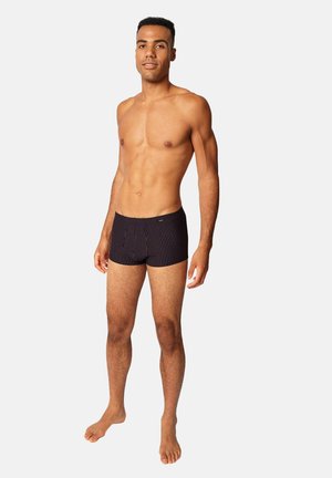 Mann mit mittlerem Hautton, trägt schwarze, schwarz gestreifte Boxer-Shorts, steht barfuß vor hellem, einfarbigem Hintergrund.