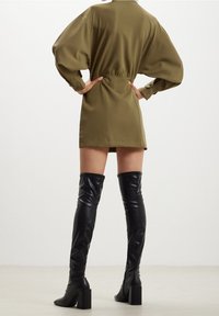 Donna che indossa un mini abito a maniche lunghe verde oliva con maniche a palloncino e stivali in pelle neri con tacco al ginocchio, in piedi con le mani sui fianchi.