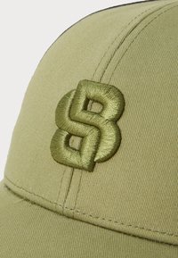 Oliven grønn caps med teksturert stoff, med et fremtredende brodert logo. Skjermen stikker fremover, med synlige sømdetaljer.