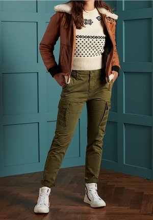 Žena v hnědém kabátu s beránkem, bílém svetr s vzorem, olivové kalhoty typu cargo a bílé vysoké tenisky, stojící před tealovým zdobeným panelem.