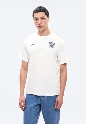 Joven con cabello corto, vestido con camiseta blanca de fútbol de Inglaterra y jeans azules, sobre un fondo liso.