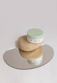 Barattolo con coperchio verde etichettato "Patch per gli occhi antiossidanti con caffeina e tè verde" sopra una pietra beige liscia, riflesso su un vassoio ovale argentato.