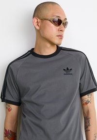 Gray cotton T-shirt with black trim, featuring a small black Adidas logo and three black stripes on sleeves. Slim-fit design, smooth texture.