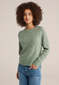 Maglione verde lavorato a maglia con un motivo a onde testurizzato e piccoli fori decorativi. Maniche lunghe e polsini a coste, abbinato a jeans blu chiaro.